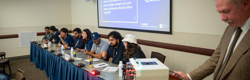 Participants sit at a table with buzzers with the moderator standing near to ask quiz questions