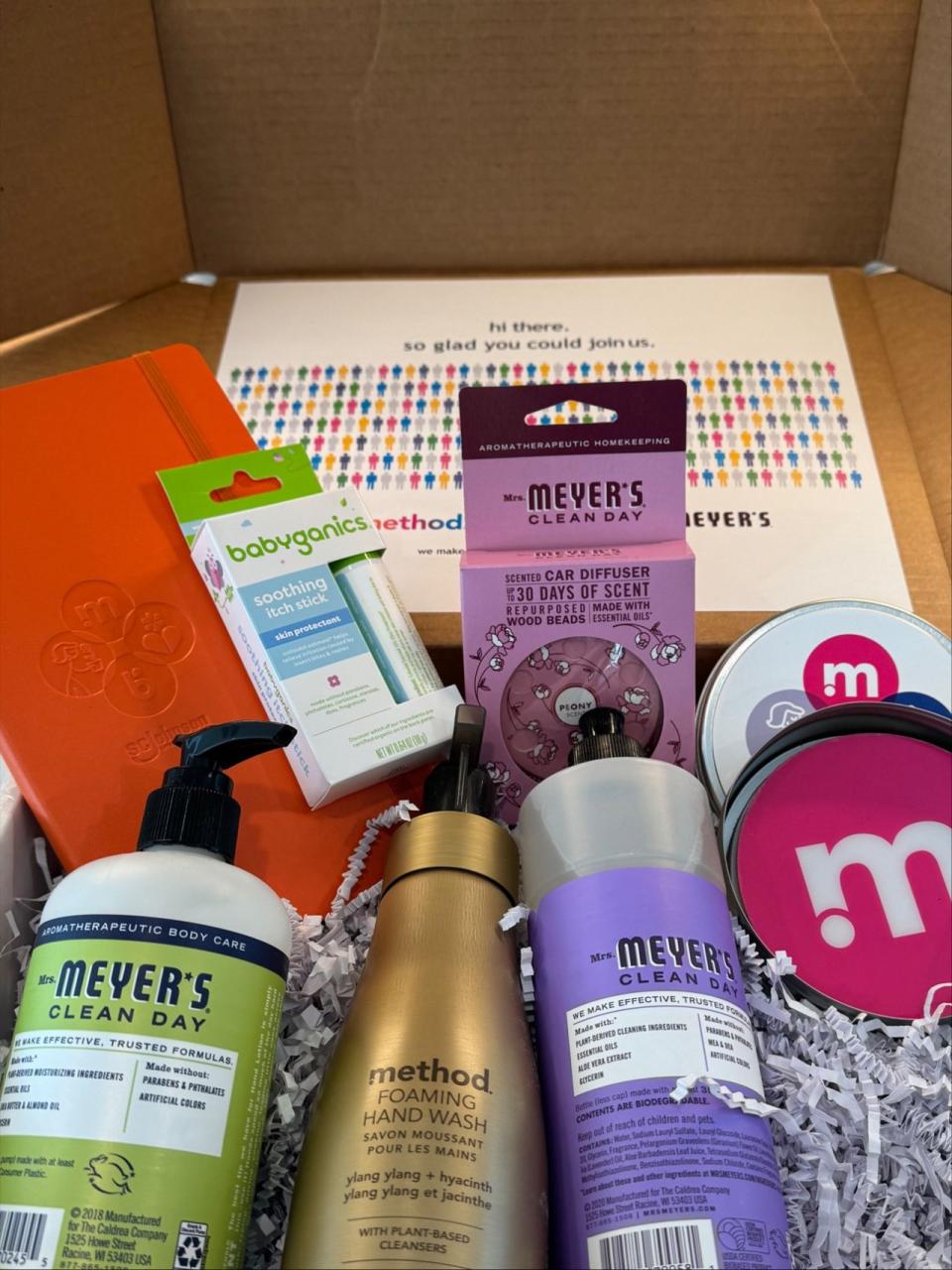 A box filled with various personal care products, including Mrs. Meyer’s Clean Day hand lotion and car diffuser, Method foaming hand wash, a Babyganics soothing itch stick, an orange notebook, and Method and Mrs. Meyer's branded items. The background features a card with colorful icons of people and a message reading 'Hi there, so glad you could join us.